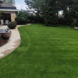 MateoGarden - Zadbany ogród z równo przystrzyżonym trawnikiem w paski, graniczący z kamienną ścieżką i elementami architektury ogrodowej. W tle widoczne drzewa i budynek.