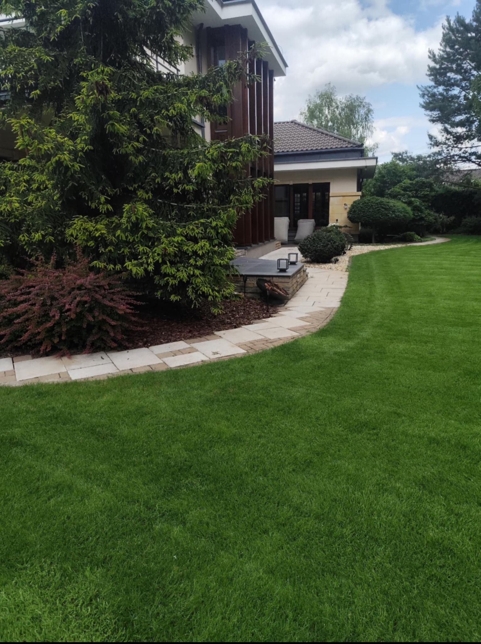 Zadbany ogród z równo przystrzyżonym trawnikiem, ozdobnymi krzewami i ścieżką z kostki brukowej prowadzącą wzdłuż nowoczesnego domu z elementami drewnianymi.