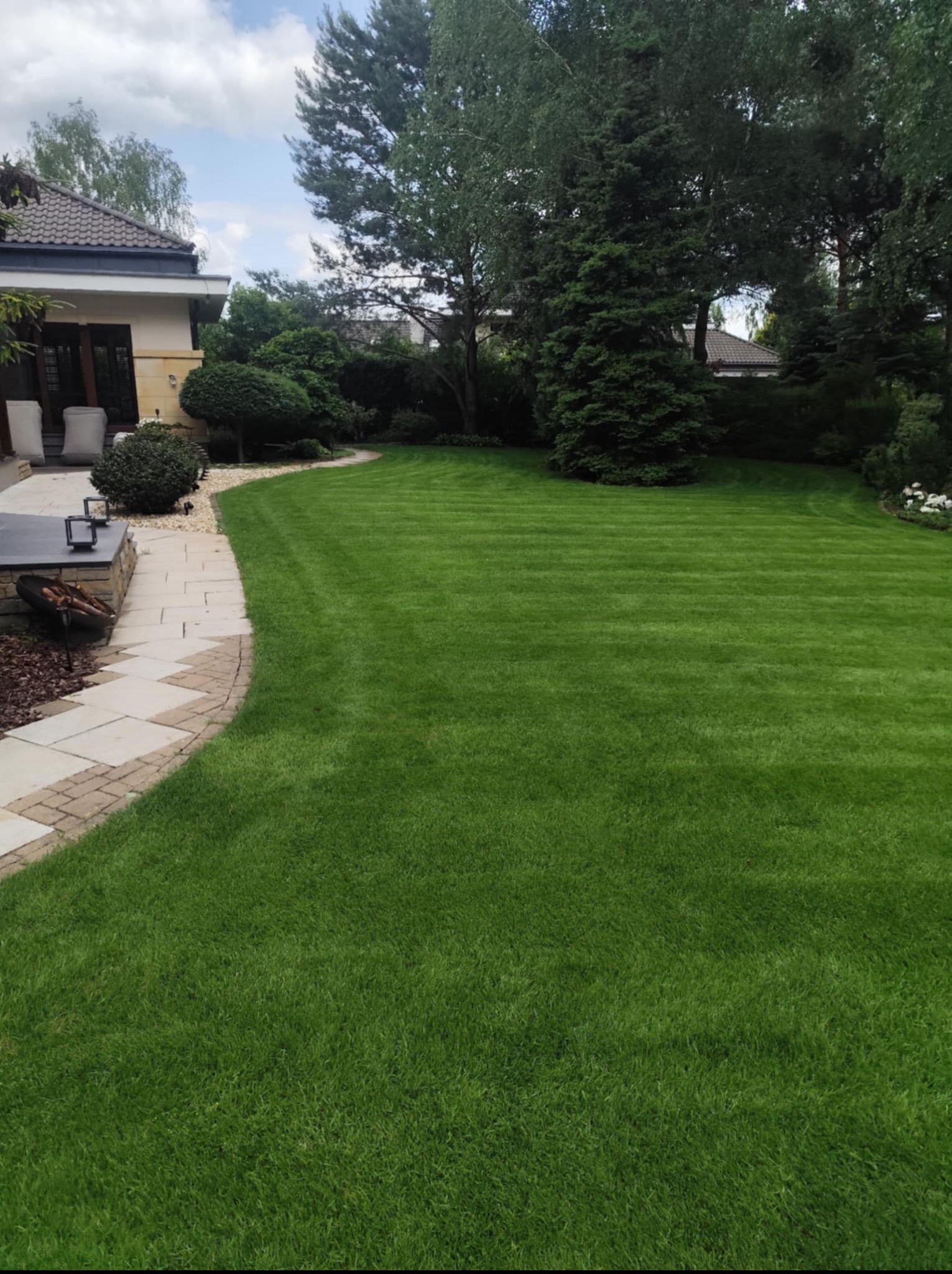 Zadbany ogród z równo przystrzyżonym trawnikiem w paski, graniczący z kamienną ścieżką i elementami architektury ogrodowej. W tle widoczne drzewa i budynek.