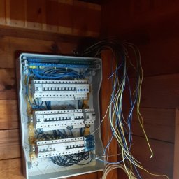 AT Urządzenia dla Domu - Rozdzielnica elektryczna z wystającymi przewodami na drewnianej ścianie. Widoczne bezpieczniki i złącza. Przewody w kolorach żółtym, niebieskim i czarnym.