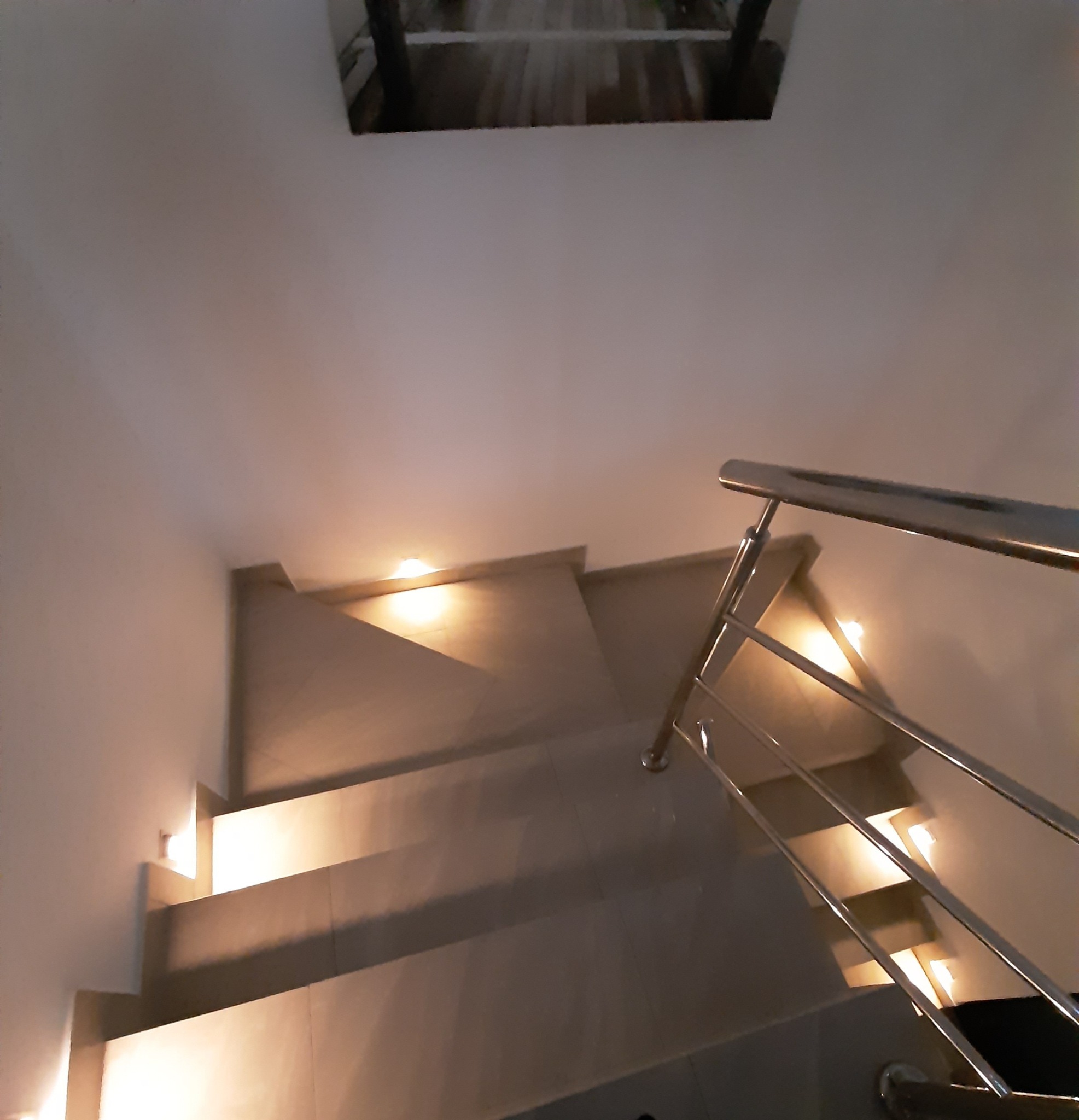 Schody z podświetleniem LED w ścianie i chromowaną balustradą, widok z góry. Na ścianie obraz. Ciepłe światło.