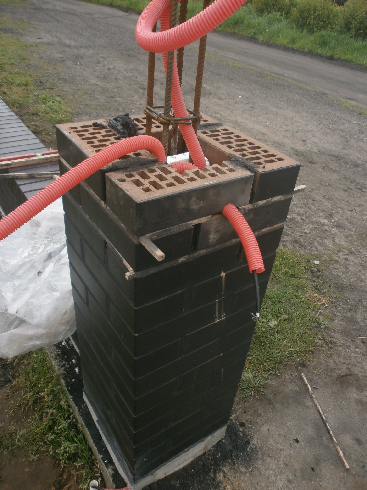 Budowa słupka z cegieł klinkierowych z peszlami na kable, w trakcie prac instalacyjnych. Widoczne zbrojenie i elementy konstrukcyjne.