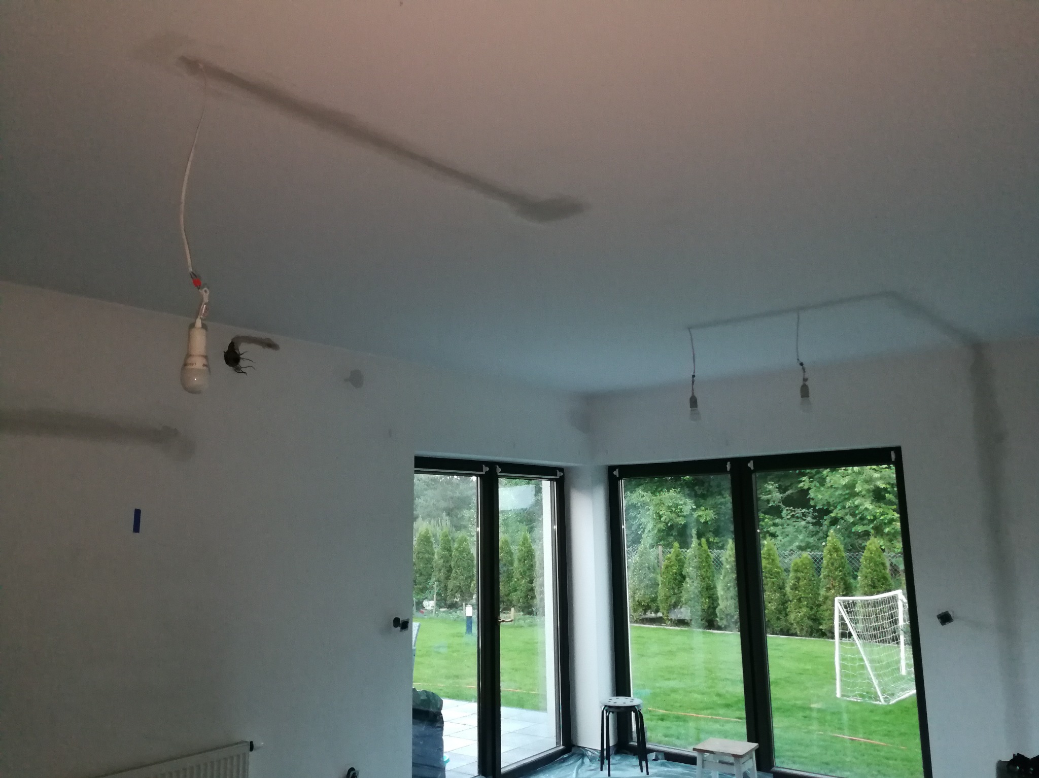 Wnętrze w trakcie prac wykończeniowych: widoczne przewody i żarówki zwisające z sufitu, przygotowane do montażu oświetlenia, duże okna z widokiem na ogród.