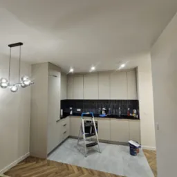 Jasna kuchnia po remoncie z drabinką malarską, widok na świeżo pomalowane ściany i nowoczesne oświetlenie. Podłoga częściowo zabezpieczona taśmą malarską.