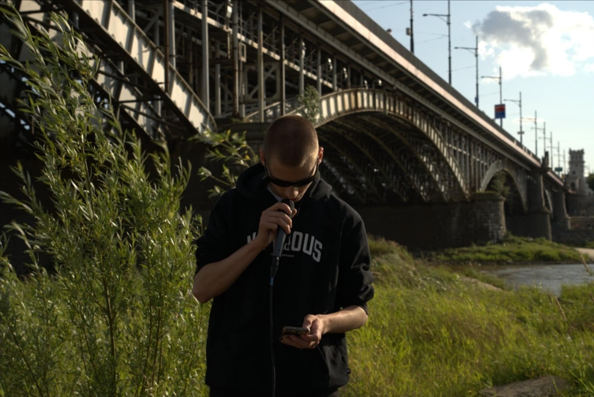 Młody mężczyzna w bluzie z kapturem, stojący na tle mostu i rzeki, trzyma mikrofon i telefon, prawdopodobnie podczas występu lub nagrania.