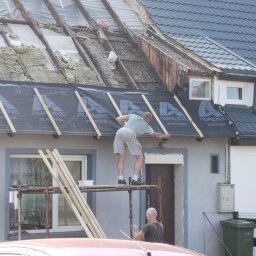 KOT-BUD - Prace dekarskie w toku: dekarz montuje membranę dachową na konstrukcji dachu jednorodzinnego domu. Część dachu z izolacją, widoczne rusztowanie i drabina.