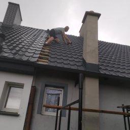 KOT-BUD - Dekarz na dachu z czarnej dachówki, montuje elementy przy kominie. Widoczne rusztowanie i okna budynku. Pochmurne niebo w tle.