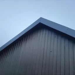 Fragment dachu z blachy trapezowej w kolorze grafitowym, widoczny od dołu na tle jasnego nieba. Szczyt dachu z obróbkami blacharskimi.