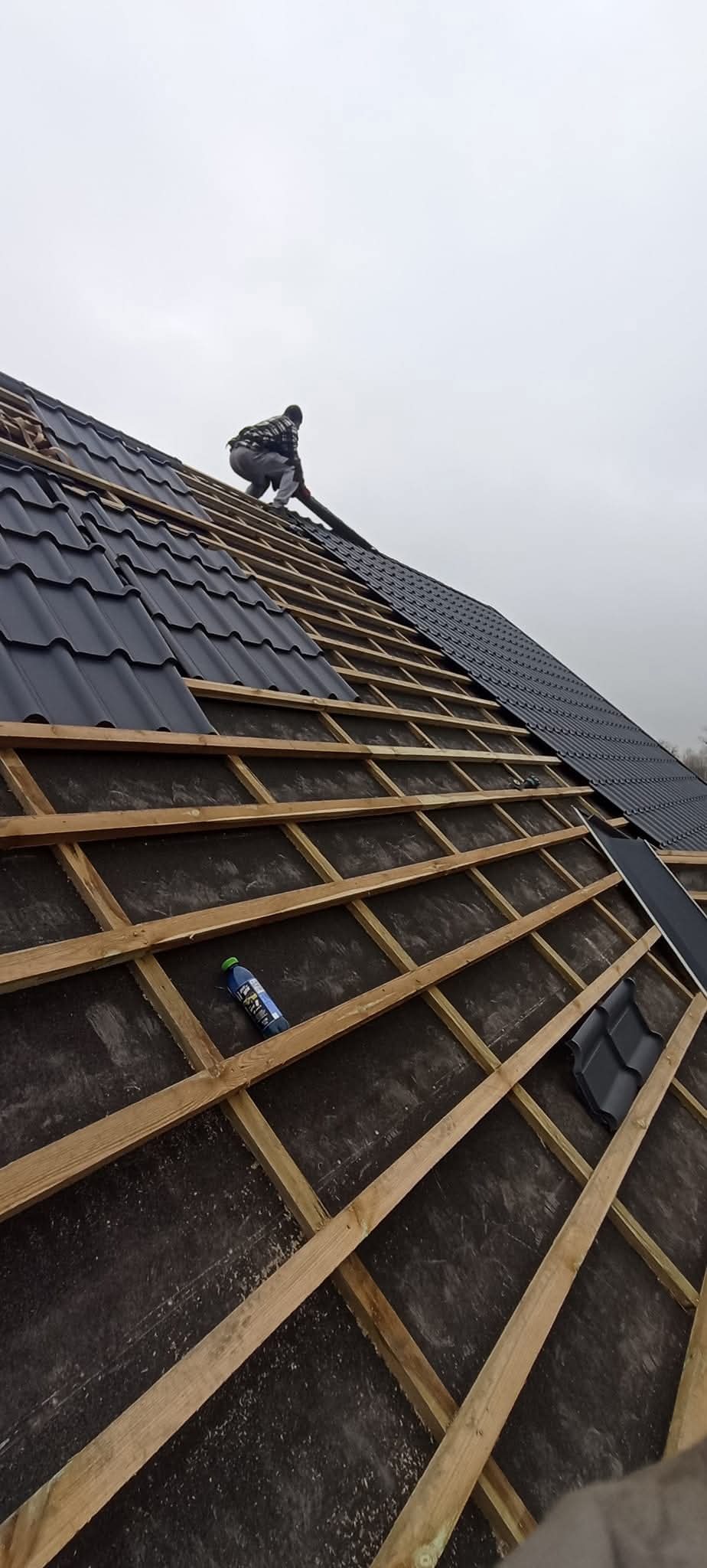 Montaż czarnej blachodachówki na drewnianej konstrukcji dachu; widoczny dekarz w trakcie pracy i butelka wody na folii dachowej. Praca na wysokości.