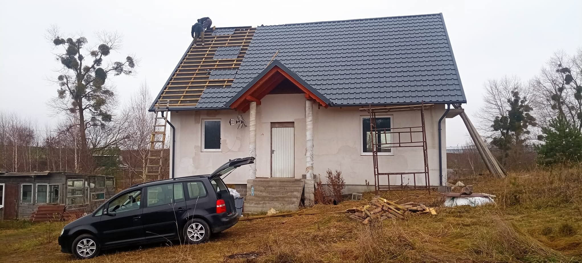 Dom jednorodzinny w trakcie krycia dachu szarą dachówką. Widoczni dekarze na dachu, rusztowanie, drabina i samochód z otwartym bagażnikiem. Jesienna sceneria.