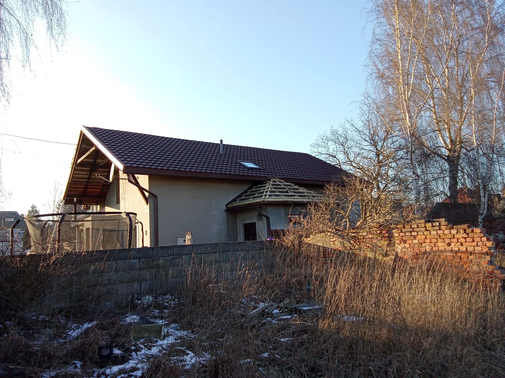 Dom z brązowym dachem z dachówki, fragment dachu w trakcie budowy, widok zza kamiennego muru i wysokiej trawy.