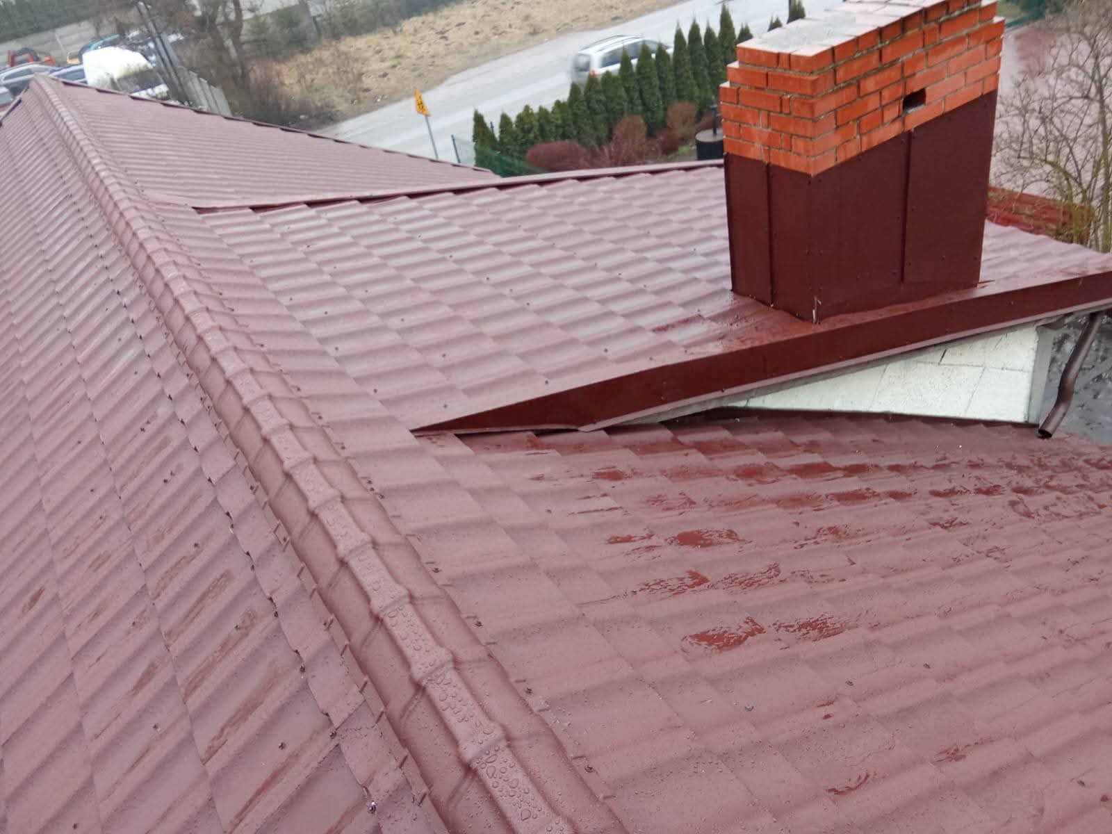 Czerwony dach z blachodachówki z ceglanym kominem i rynną, widok z góry po deszczu. Widoczne krople wody na powierzchni. Dach po renowacji.