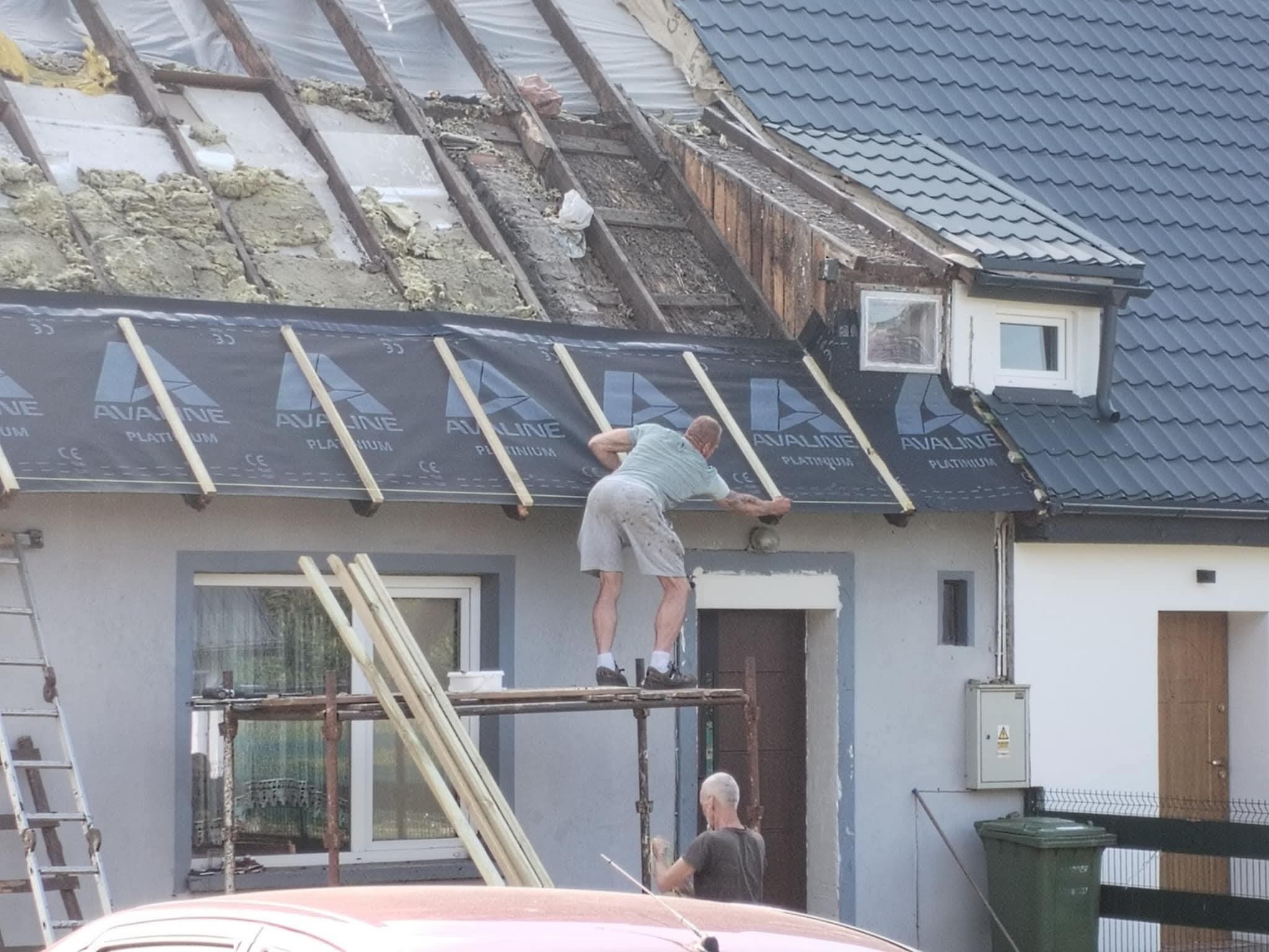 Prace dekarskie w toku: dekarz montuje membranę dachową na konstrukcji dachu jednorodzinnego domu. Część dachu z izolacją, widoczne rusztowanie i drabina.