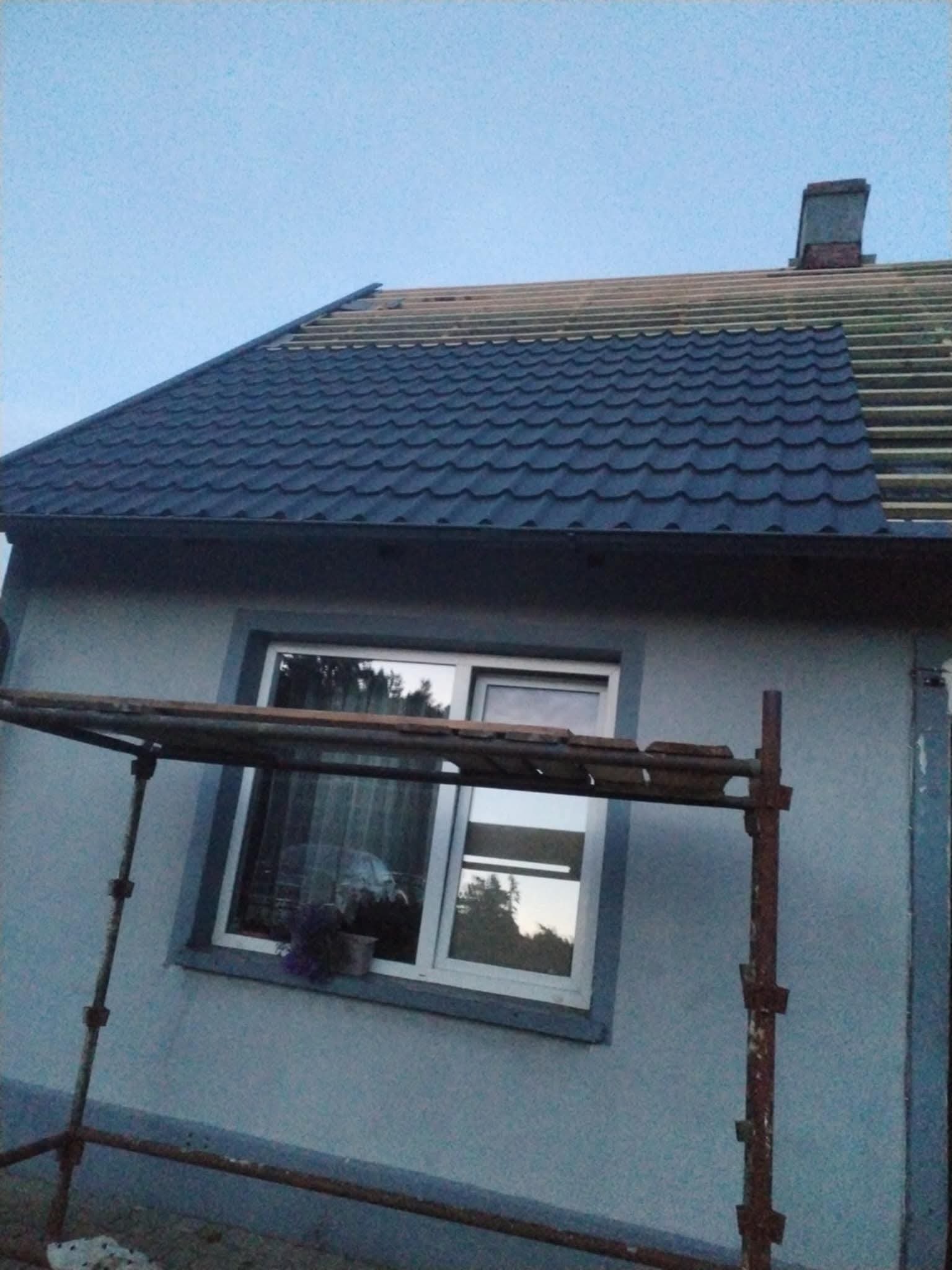 Częściowo pokryty dach z ciemnej blachodachówki, widoczne łaty i komin. Na pierwszym planie rusztowanie przy oknie domu.