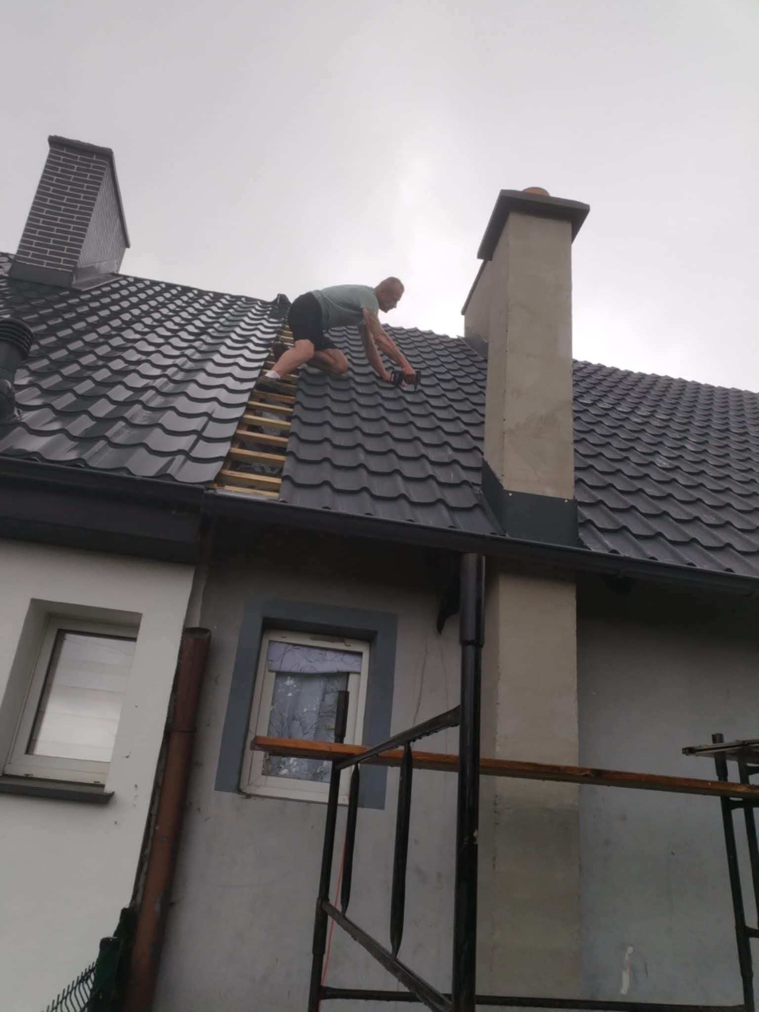 Dekarz na dachu z czarnej dachówki, montuje elementy przy kominie. Widoczne rusztowanie i okna budynku. Pochmurne niebo w tle.