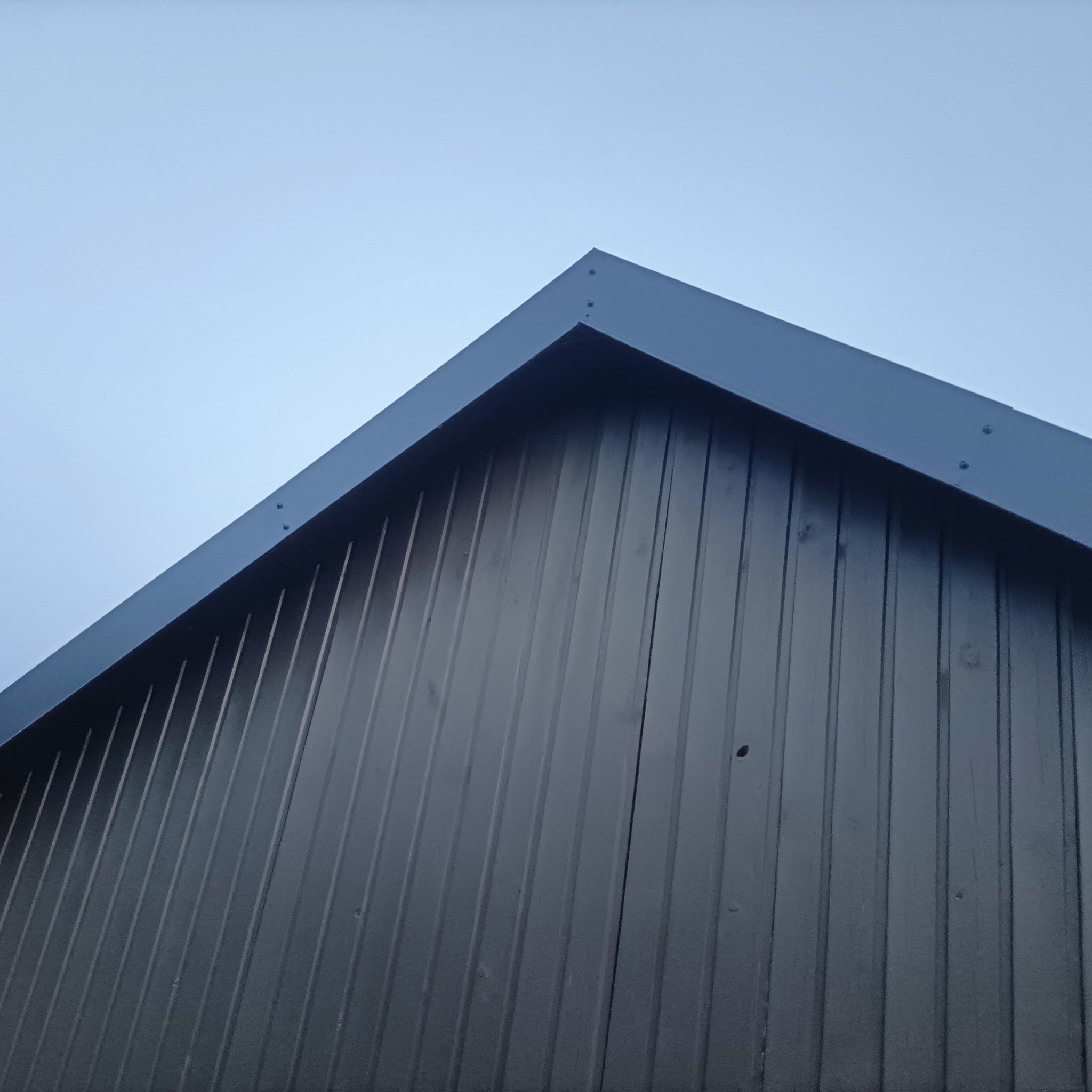 Fragment dachu z blachy trapezowej w kolorze grafitowym, widoczny od dołu na tle jasnego nieba. Szczyt dachu z obróbkami blacharskimi.