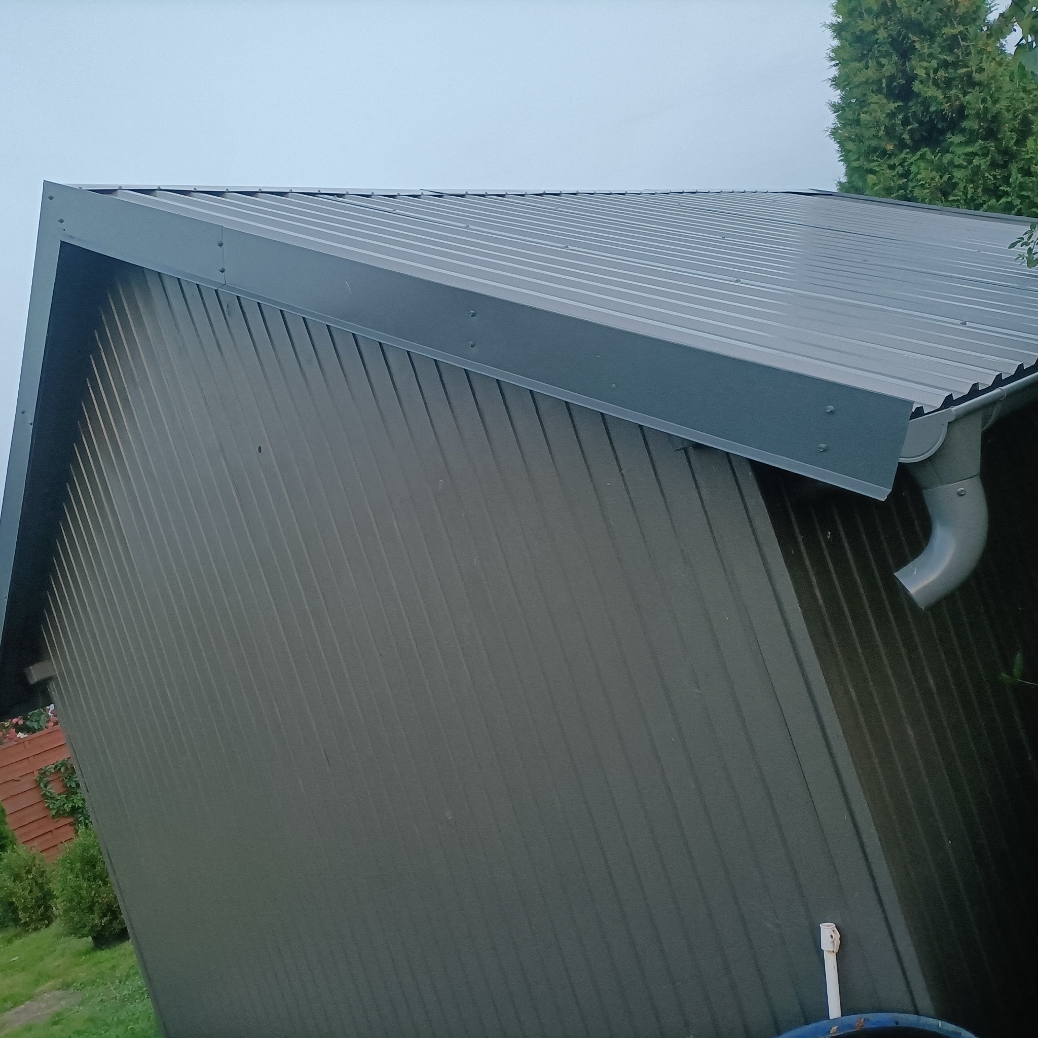 Szary dach z blachy trapezowej z zamontowaną rynną, widok z dołu, na tle zieleni i błękitnego nieba. Detal dachu z blachy.