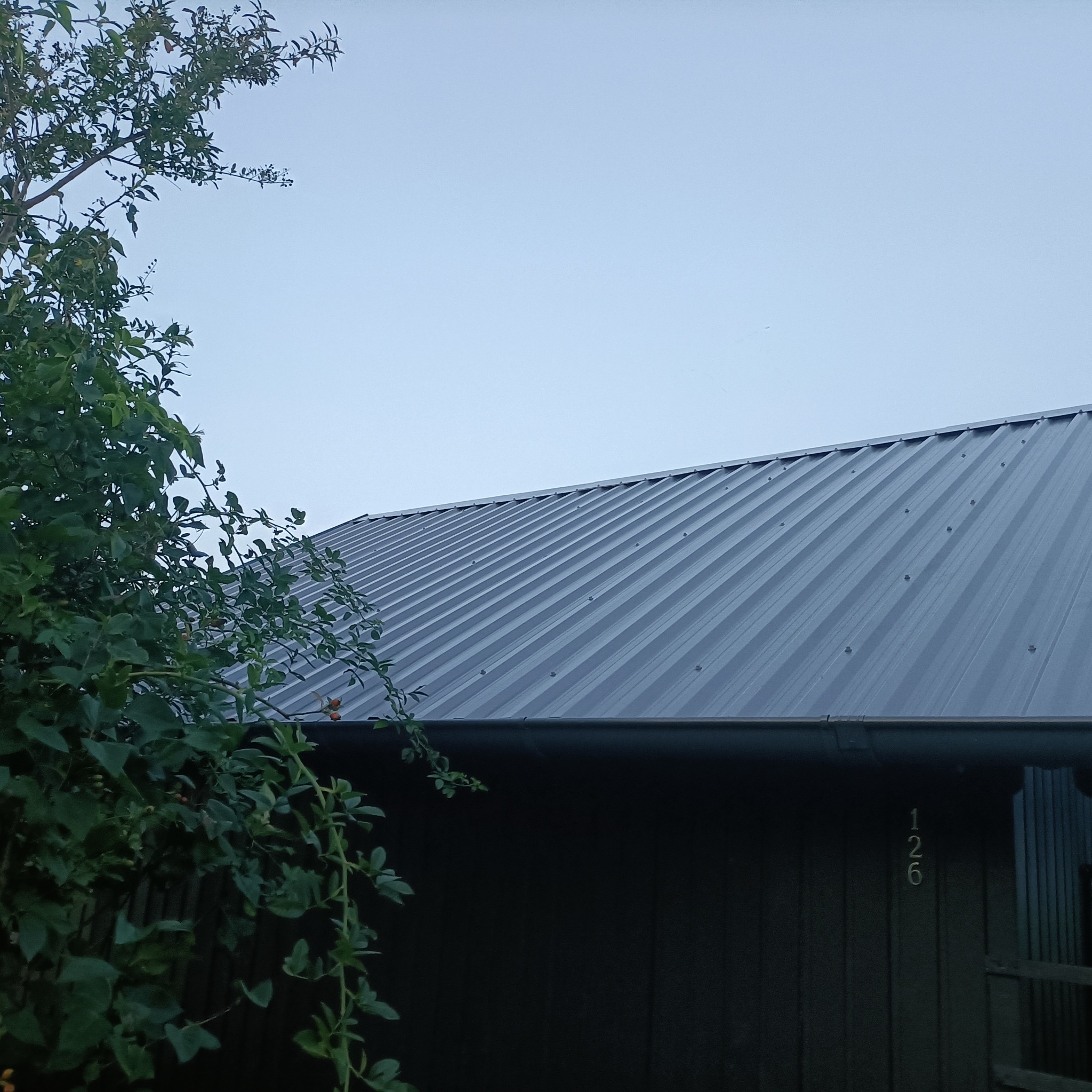 Szary dach z blachy trapezowej, widoczny z dołu, z zielenią drzewa po lewej stronie i numerem domu 126 na ścianie budynku.