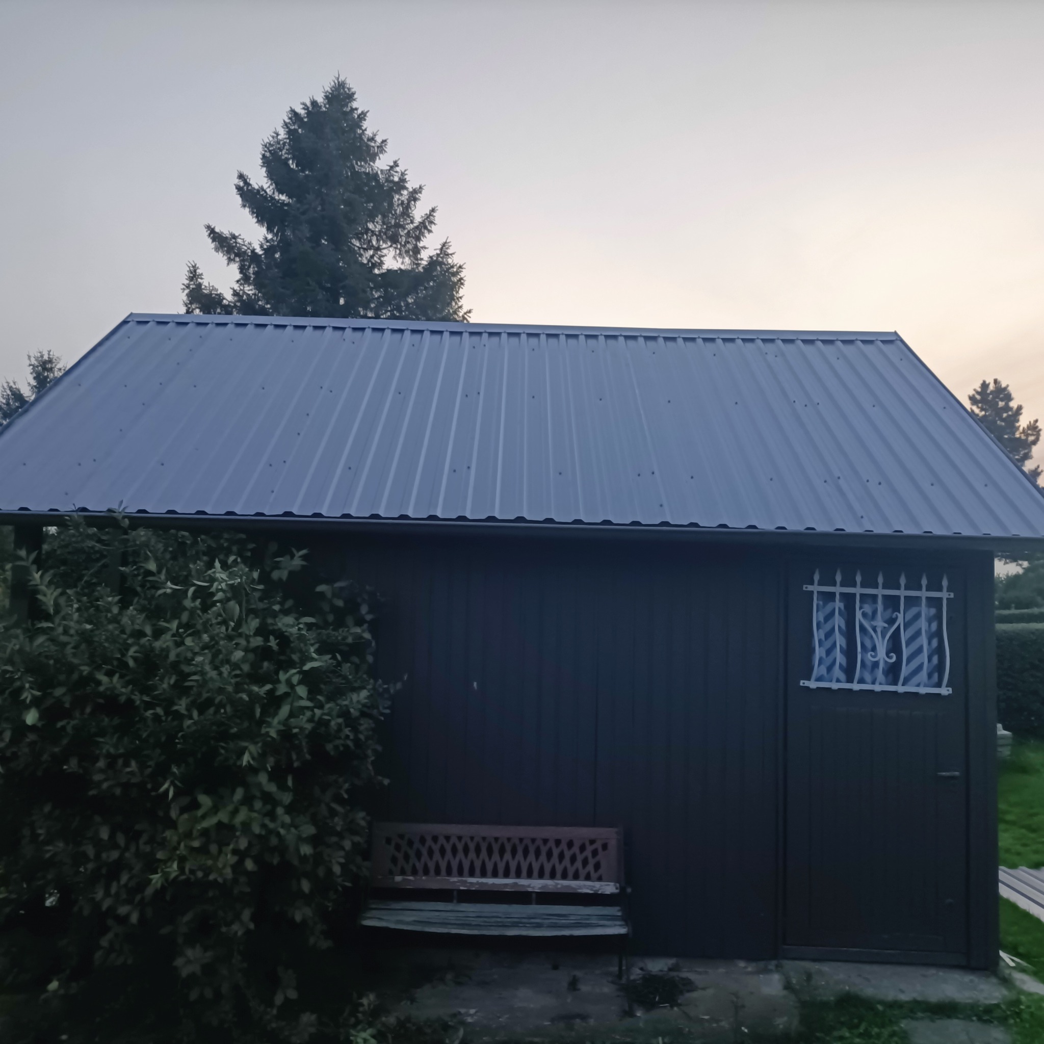 Metalowy dach na budynku gospodarczym w Skarszewach. Nowe krycie dachu z blachy trapezowej, widoczna boczna ściana budynku i zieleń w tle.