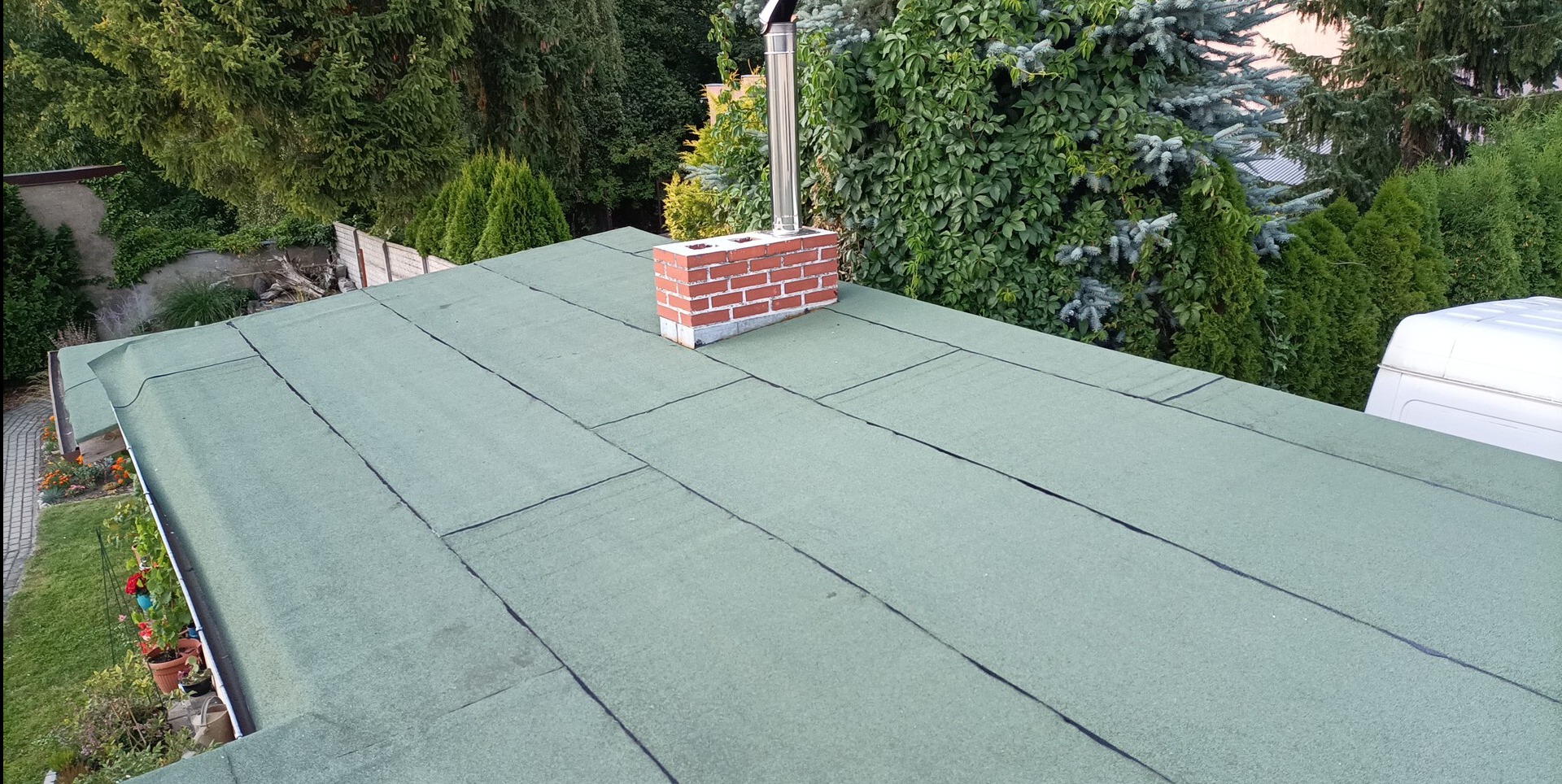 Zielony dach pokryty papą termozgrzewalną z kominem z cegły i metalowym wkładem kominowym, otoczony zielenią. Widok z góry.