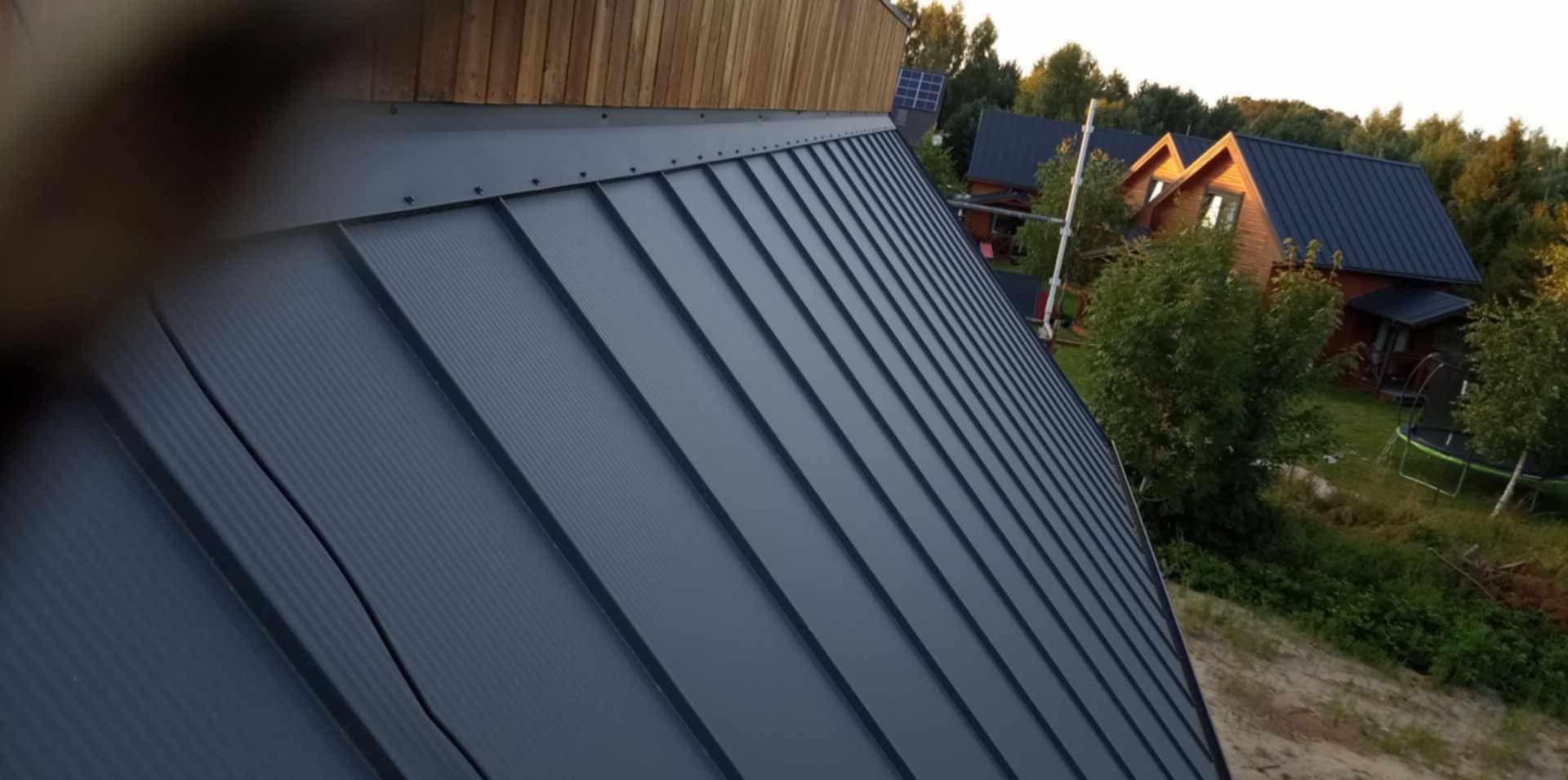 Szary dach z blachy na domu z drewnianą elewacją, widok z góry, na tle drzew i innych domów. Nowoczesne pokrycie dachowe.