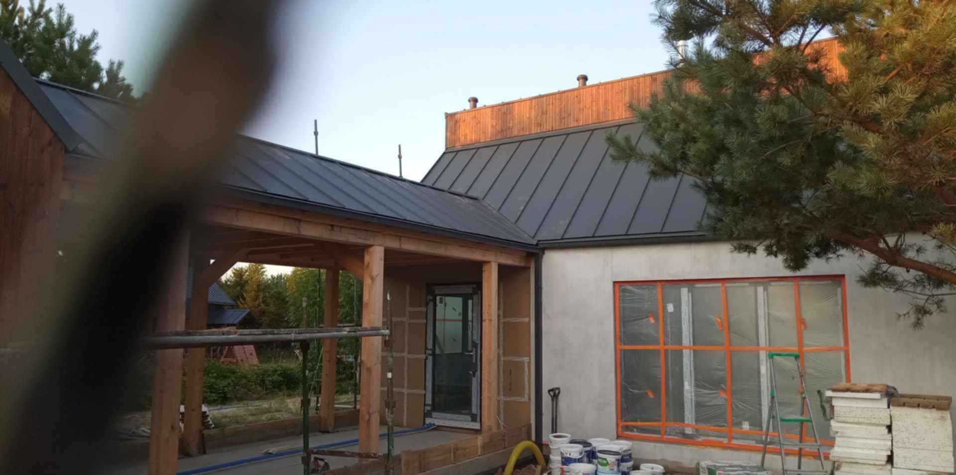 Nowoczesny dom z grafitowym dachem z blachy na rąbek stojący, drewnianą konstrukcją ganku i oknem zabezpieczonym folią malarską. Częściowo otynkowana elewacja.