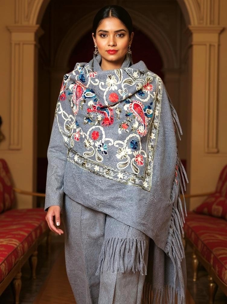 Modelka prezentuje szary, wełniany komplet z frędzlami i bogato haftowanym szalem w kwiatowe wzory, stojąc na tle klasycznego wnętrza.