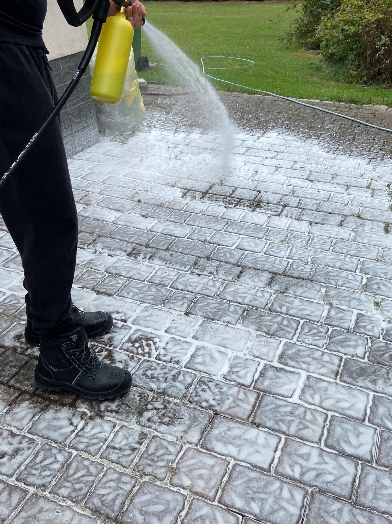 Czyszczenie kostki brukowej myjką ciśnieniową z żółtym pojemnikiem na detergent. Widoczne czarne buty pracownika i fragment węża wysokociśnieniowego.