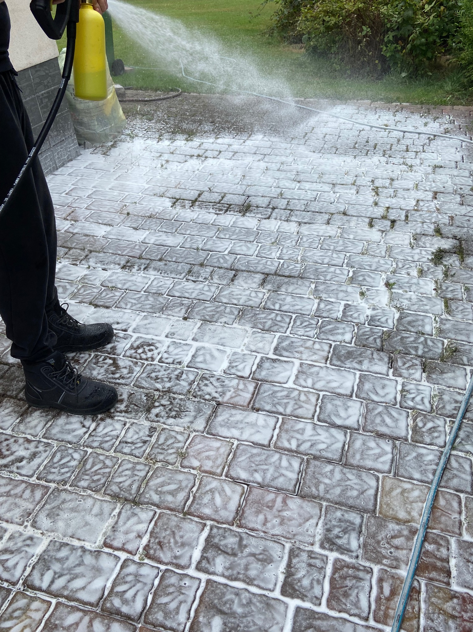 Czyszczenie kostki brukowej myjką ciśnieniową; widoczne buty robocze, spieniona powierzchnia i strumień wody. Prace w toku na tle zieleni.