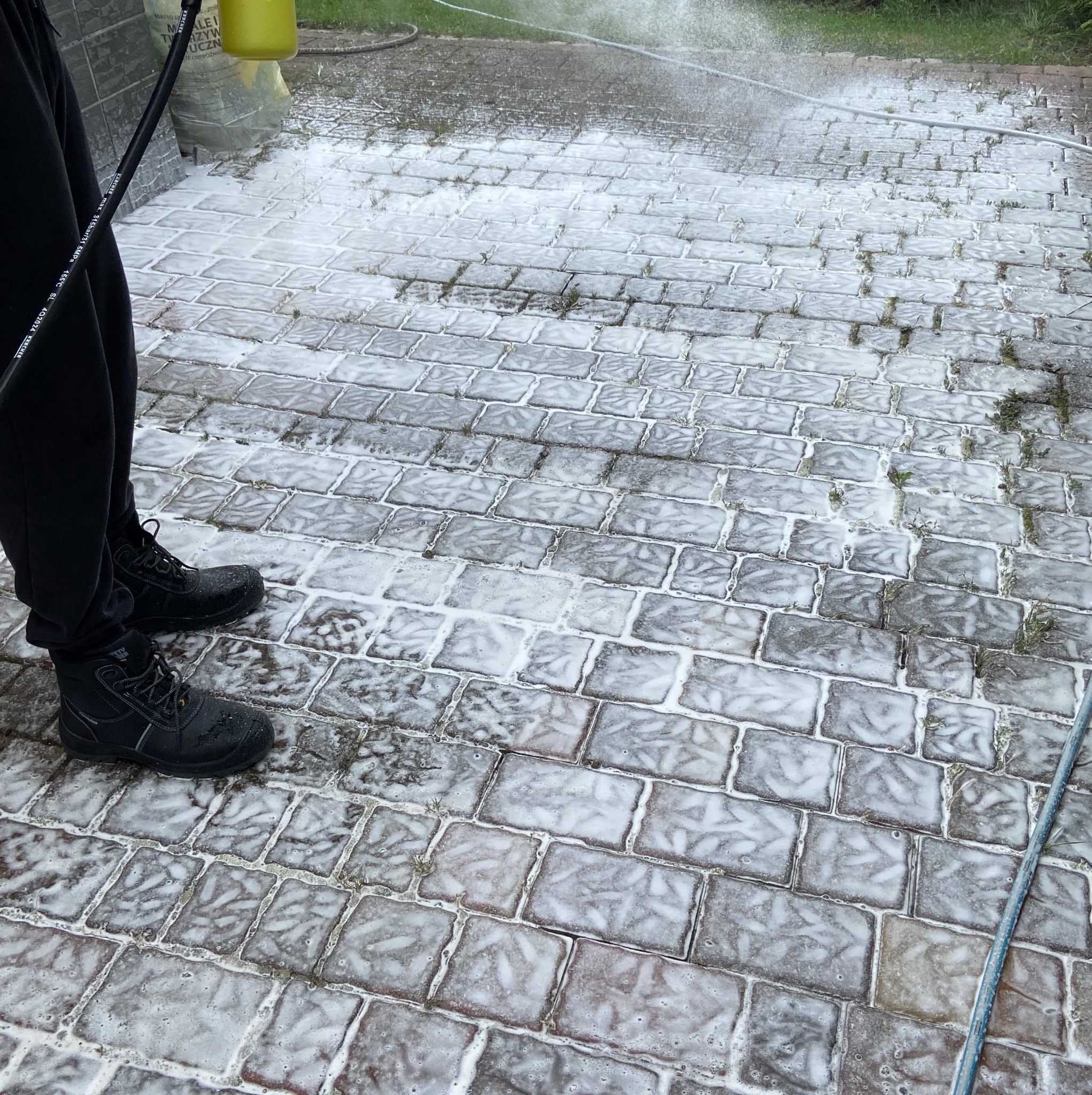 Czyszczenie kostki brukowej z użyciem myjki ciśnieniowej. Piana pokrywa szare kostki, widoczne buty pracownika i wąż myjki. Częściowo umyta powierzchnia.
