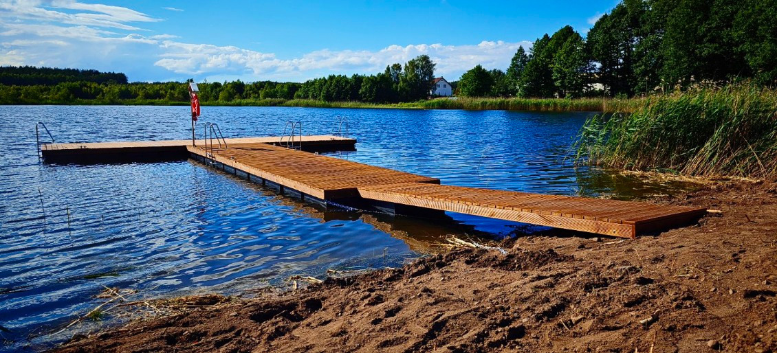 Drewniany pomost z drabinkami na jeziorze w słoneczny dzień. Czerwona boja na maszcie. W tle las i biały budynek.
