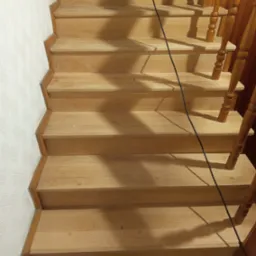 Jasne, drewniane schody z balustradą, widok z g&oacute;ry. Kabel od urządzenia do obr&oacute;bki drewna leży na stopniach. Minimalistyczny wz&oacute;r na ścianie.