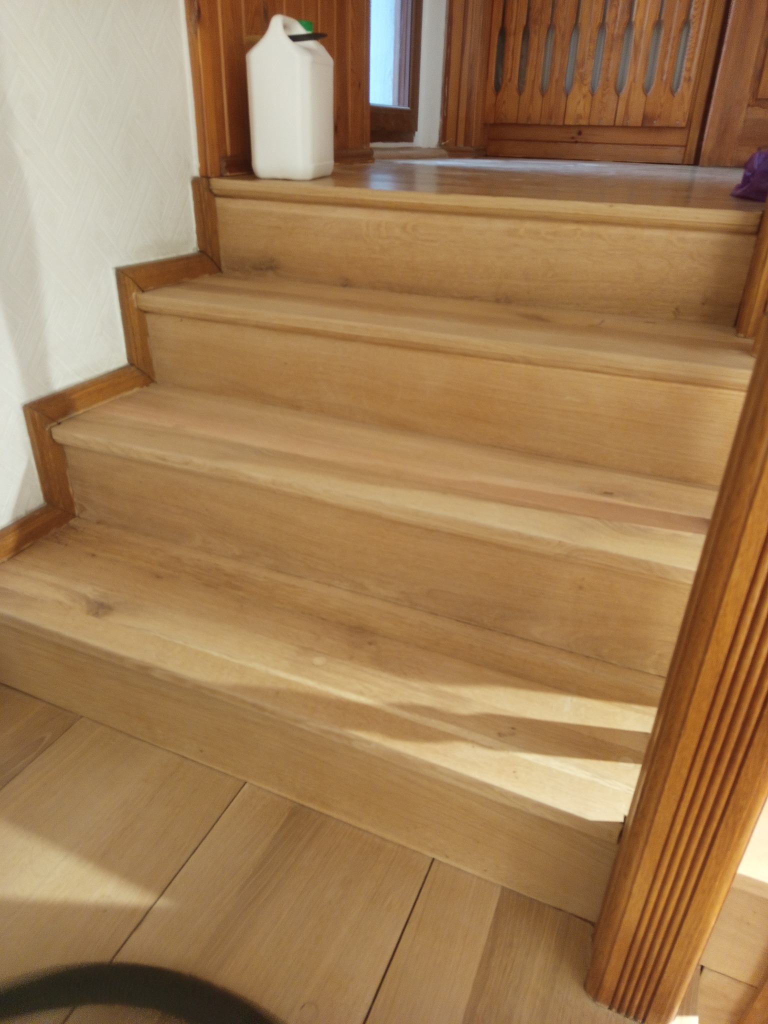 Jasne, drewniane schody z widocznym słojem drewna, prowadzące do góry. Biały pojemnik na górnym stopniu. Parkiet na podłodze. Element dekoracyjny.