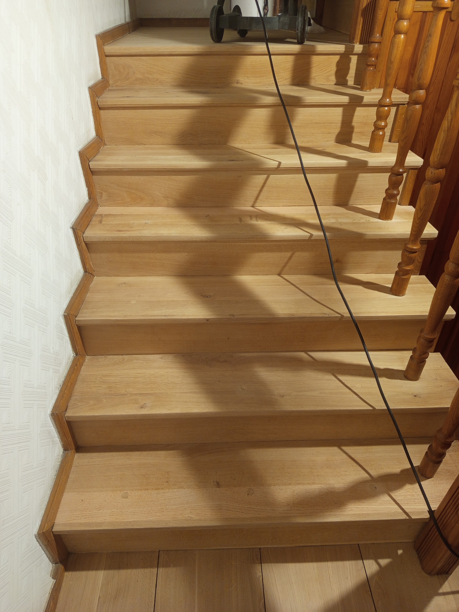 Jasne, drewniane schody z balustradą, widok z góry. Kabel od urządzenia do obróbki drewna leży na stopniach. Minimalistyczny wzór na ścianie.