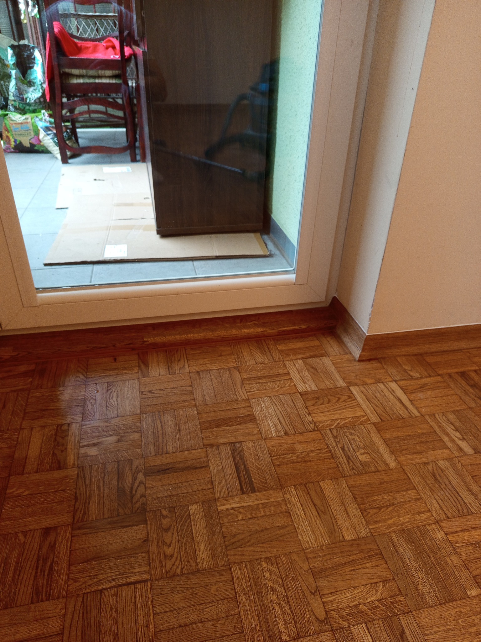 Parkiet z drewna ułożony w kwadraty, widok na próg drzwi balkonowych. Podłoga wykończona listwami przypodłogowymi. Część podłogi zakryta kartonem.