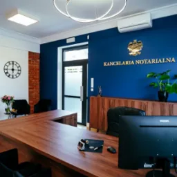 Eleganckie wnętrze kancelarii: drewniane biurko, niebieska ściana z napisem 'Kancelaria Notarialna', ceglana ściana, nowoczesne oświetlenie i elementy dekoracyjne.