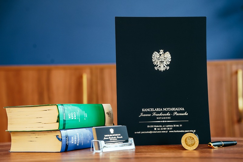 Kancelaria notarialna: akta, wizytówka, kodeksy, moneta i pióro na drewnianym biurku, na tle ściany w dwóch odcieniach niebieskiego i brązu.