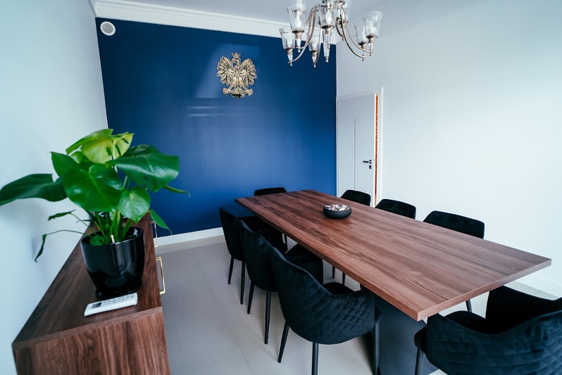 Sala konferencyjna z długim drewnianym stołem, czarnymi krzesłami i godłem Polski na niebieskiej ścianie. Roślina doniczkowa na komodzie.