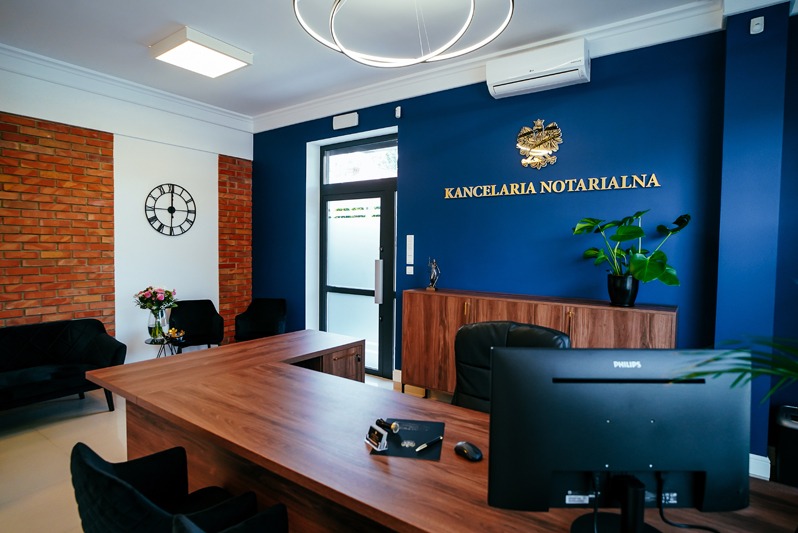 Eleganckie wnętrze kancelarii: drewniane biurko, niebieska ściana z napisem 'Kancelaria Notarialna', ceglana ściana, nowoczesne oświetlenie i elementy dekoracyjne.