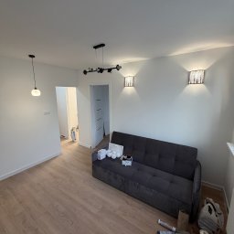 DAWID GRUSE DG USŁUGI - Jasne wnętrze po remoncie: drewniana podłoga, szara sofa, białe ściany z nowoczesnym oświetleniem. Widoczne narzędzia malarskie sugerują świeże wykończenie.