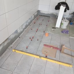 DAWID GRUSE DG USŁUGI - Układanie szarych płytek podłogowych w łazience z użyciem poziomicy i systemu poziomowania. Widoczne narzędzia i materiały budowlane. Ściany wyłożone jasnymi płytkami.
