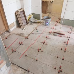 DAWID GRUSE DG USŁUGI - Układanie płytek podłogowych w łazience z użyciem systemu poziomowania. Widoczne płytki, krzyżyki dystansowe, poziomica i wiadro z klejem Atlas. Prace glazurnicze w toku.