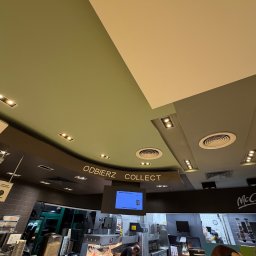 DAWID GRUSE DG USŁUGI - Wnętrze lokalu gastronomicznego z widocznym sufitem w odcieniach beżu i zieleni, oświetleniem punktowym i napisem 'ODBIERZ COLLECT'. W tle stanowisko pracy z pracownikiem.