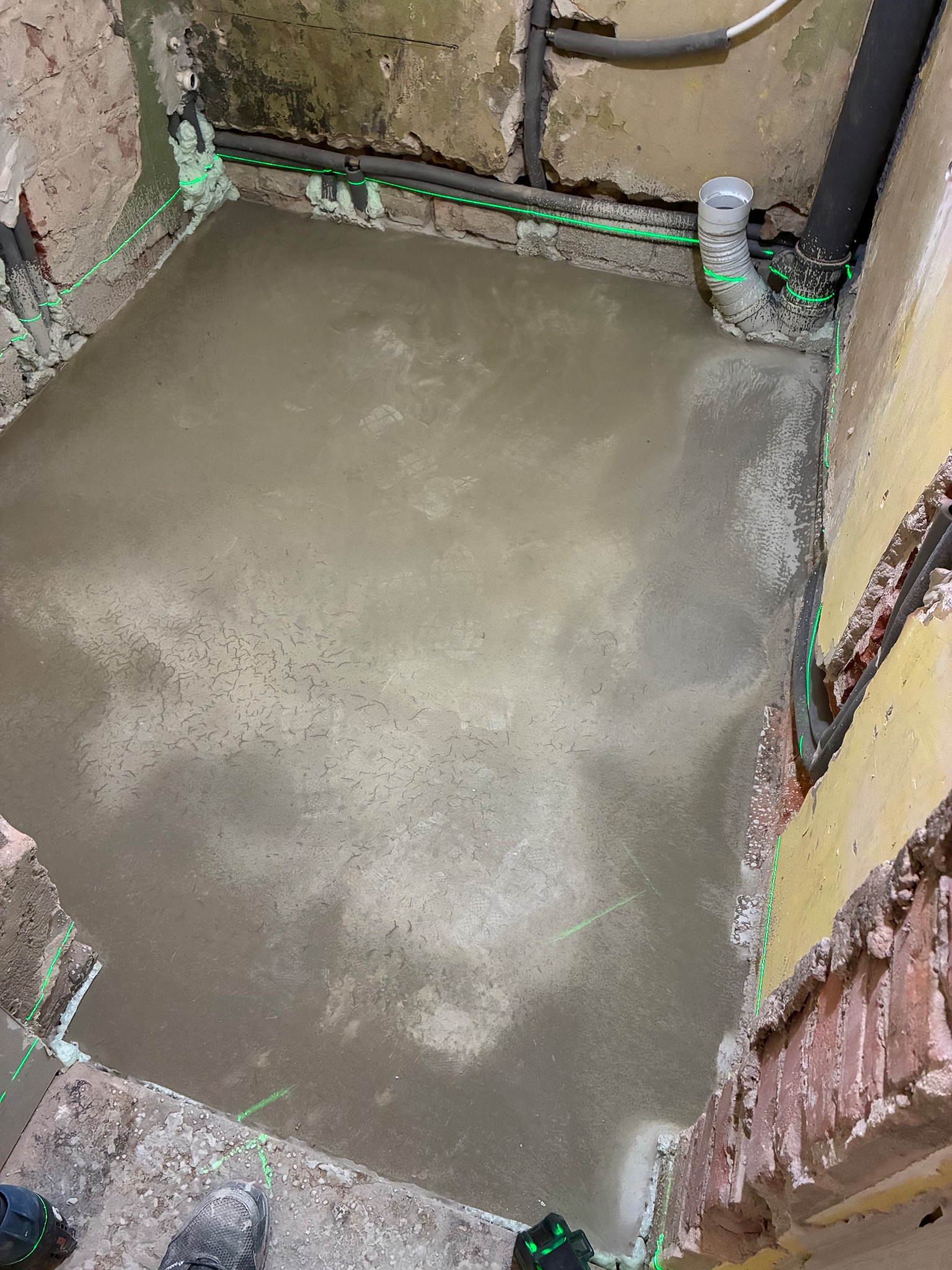 Wyrównana, świeża wylewka podłogowa w łazience, z widocznymi rurami i poziomowaniem laserowym. Ściany w trakcie remontu, częściowo otynkowane.