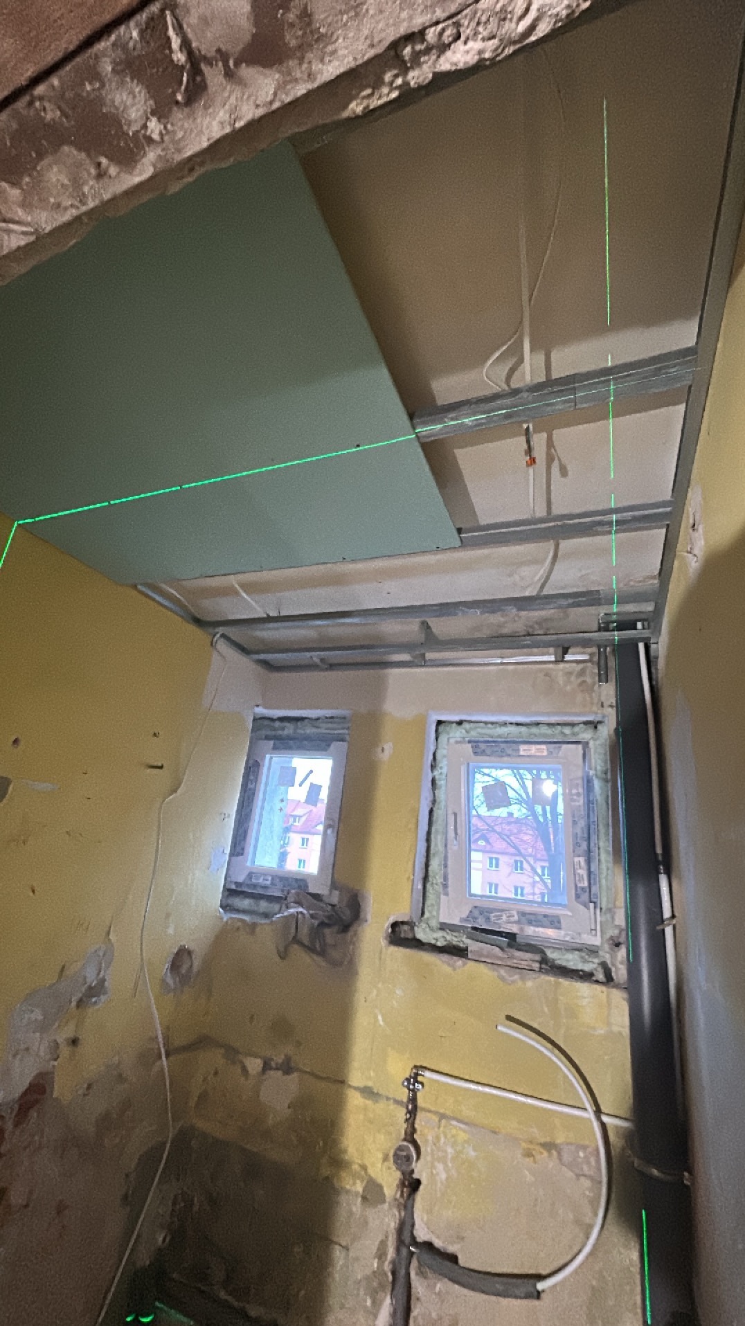 Montaż sufitu podwieszanego z płyt gipsowo-kartonowych, widoczne profile metalowe i laserowy niwelator. Ściany w trakcie remontu, dwa okna. Prace wykończeniowe.