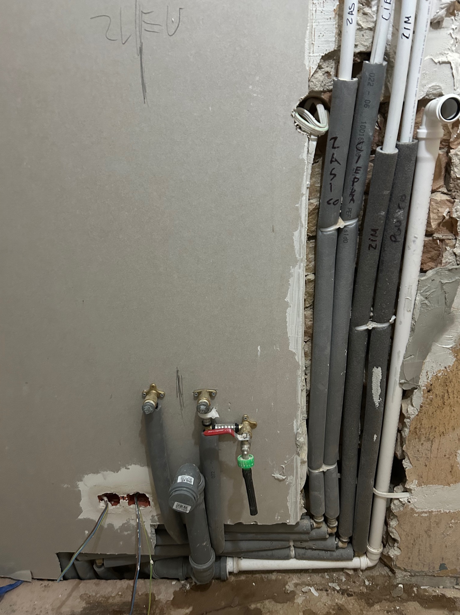 Szare rury z izolacją i białe rury wodne w ścianie podczas montażu instalacji w łazience. Widoczne przyłącza i kable elektryczne. Ściana częściowo otynkowana.