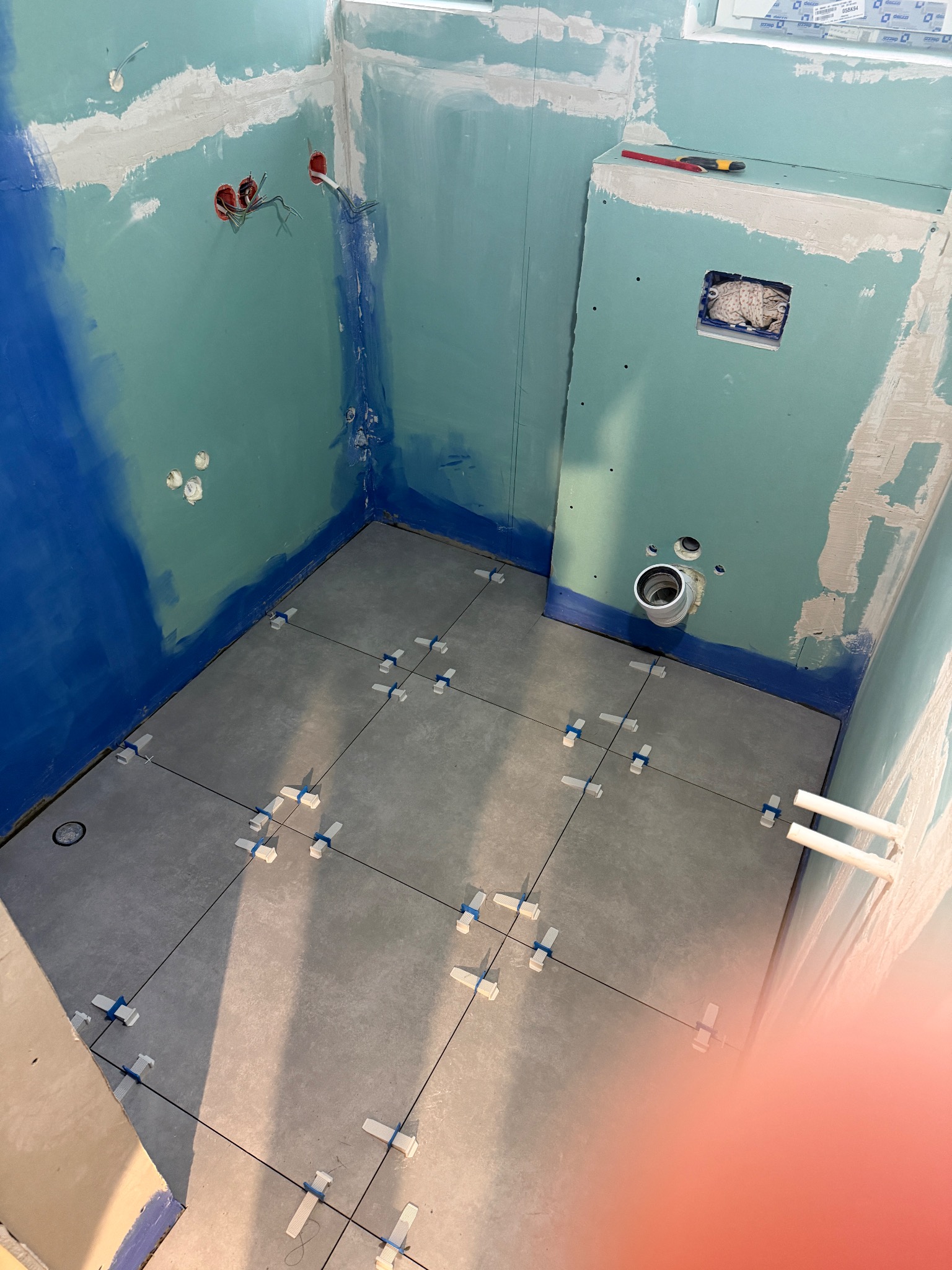 Łazienka w trakcie układania płytek podłogowych, widoczne krzyżyki dystansowe i hydroizolacja na ścianach. Przygotowane instalacje elektryczne i hydrauliczne.