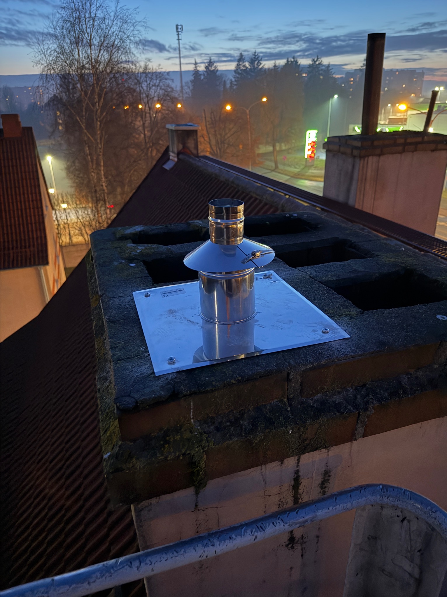 Nowy, błyszczący komin ze stali nierdzewnej zamontowany na starym, ceglanym kominie na tle wieczornego nieba i zabudowań miejskich. Widok z góry.