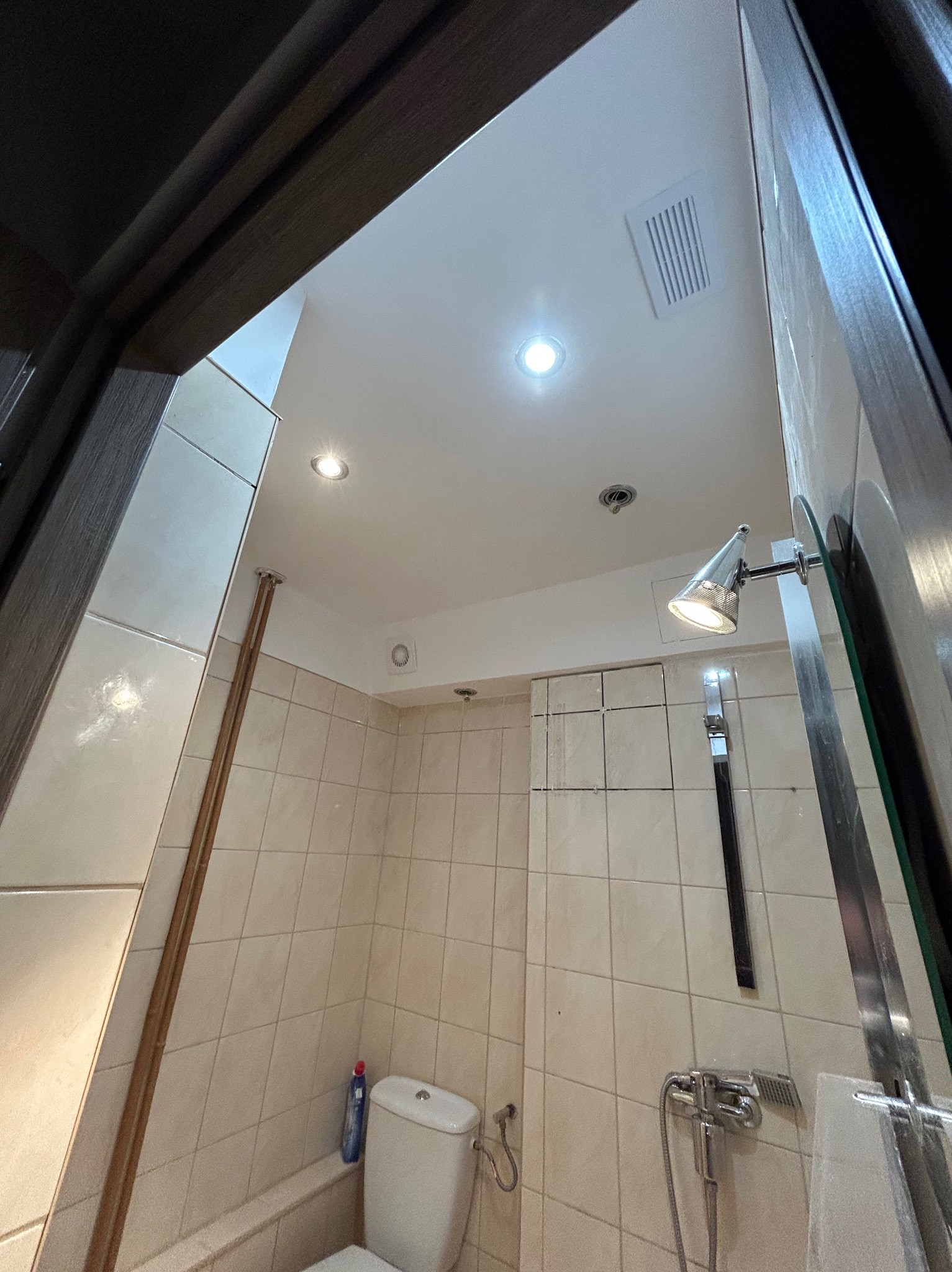 Wykończona łazienka z beżowymi płytkami, widok z dołu na sufit z oświetleniem punktowym i wentylacją. Umywalka i bateria prysznicowa w kadrze.