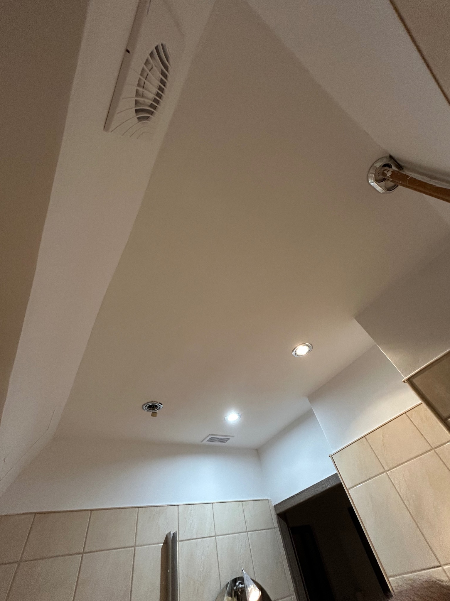 Biały sufit łazienki z kratką wentylacyjną, oświetleniem punktowym i fragmentem płytek. Ujęcie z dołu, podkreślające detale wykończenia.